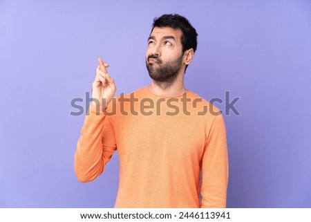 Caucasian handsome man over isolated purple background with fingers crossing and wishing the best