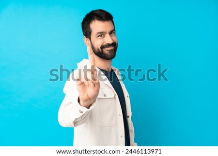 Young handsome man with white corduroy jacket over isolated blue background showing and lifting a finger
