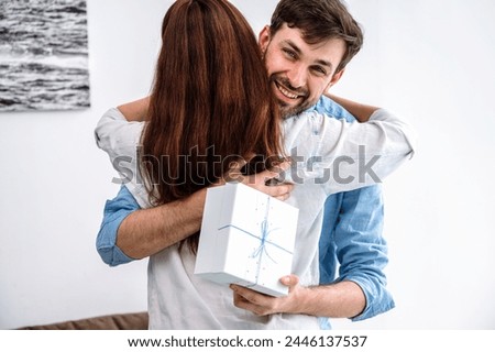 Young happy man receive gift from lovely wife, feel gratitude and hug tender her. Family couple celebrating anniversary at home and giving romantic present each other expression love and devotion Royalty-Free Stock Photo #2446137537