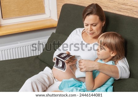 A pregnant woman in a white shirt sharing ultrasound images with her curious young daughter who examining them with great interest. A bonding moment between the sister-to-be and the unborn baby
