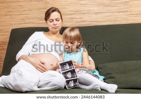 A pregnant woman in a white shirt sharing ultrasound images with her curious young daughter who examining them with great interest. A bonding moment between the sister-to-be and the unborn baby