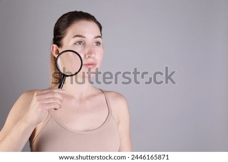 Young woman with acne problem holding magnifying glass near her skin on light grey background. Space for text
