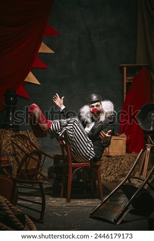 Clown in black hat, white face with red nose and striped pants performing over dark retro circus backstage background. Emotional play. Concept of circus, theater, performance, show, retro and vintage Royalty-Free Stock Photo #2446179173