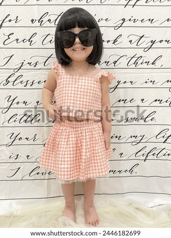 A sweet, stylish smiling little girl wearing a light orange dress, sunglasses, and a ribbon bag