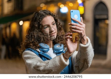 Happy teenage girl taking photography pictures of city buildings outdoors. Caucasian woman blogger using smartphone photography app making photos for social media standing on street at evening night.