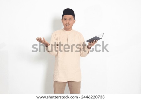 Portrait of confused Asian muslim man in koko shirt with peci difficulty understanding the contents of the book, reading a textbook. Isolated image on white background Royalty-Free Stock Photo #2446220733
