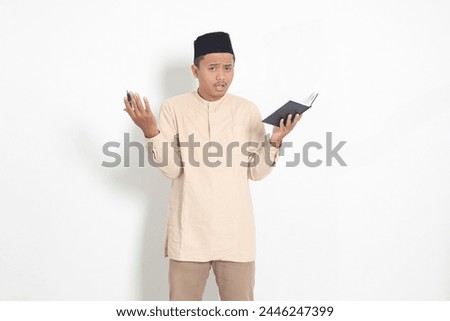 Portrait of confused Asian muslim man in koko shirt with peci difficulty understanding the contents of the book, reading a textbook. Isolated image on white background Royalty-Free Stock Photo #2446247399