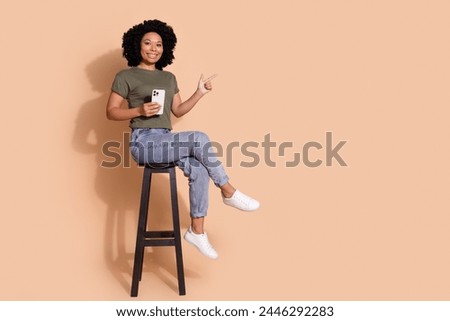 Full size photo of smart person wear t-shirt on chair hold smartphone indicating at logo empty space isolated on pastel color background