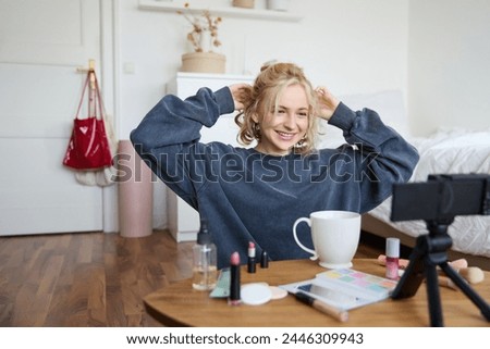 Portrait of cute blond girl, lifestyle blogger sits on floor with video camera and stabiliser, shows how to do hairstyle and makeup, does casual vlogging for social media, sits in room.