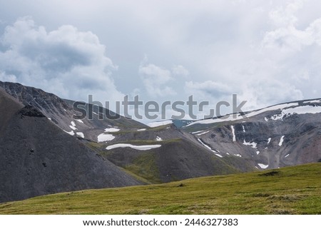 Dramatic alpine landscape with green meadow and high rocky mountain range with snowfields and ice cornices under grey cloudy sky. Shadows of clouds on large rock ridge with snow in changeable weather.