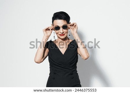 Stylish older woman in black dress and sunglasses posing in front of white wall for camera portrait Royalty-Free Stock Photo #2446375553