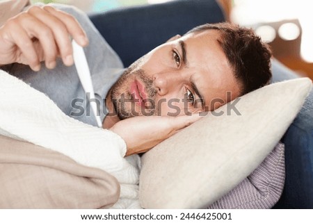 Man in couch, sick and thermometer for fever, healthcare and wellness with virus, viral infection or bacteria at home. Burnout, fatigue and health fail, monitor condition for illness with cold or flu Royalty-Free Stock Photo #2446425001