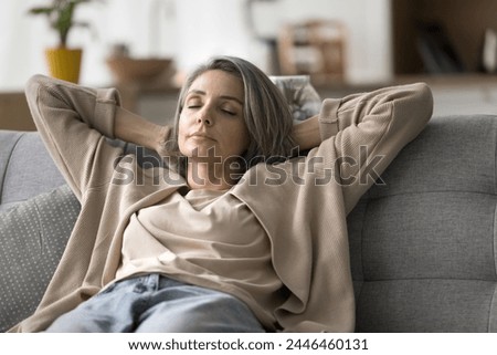 Peaceful attractive mature woman relaxing on sofa with eyes closed, lean back with hands behind head, felt asleep, relieving fatigue, breath fresh air inside modern living room, enjoy weekend at home Royalty-Free Stock Photo #2446460131