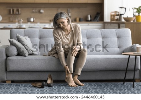 Female came home sit on sofa took off shoes, touch ankle doing self-massage of tired feet, suffers from varicose, painful feelings discomfort after long workday on heels, need rest, orthopedic insoles Royalty-Free Stock Photo #2446460145