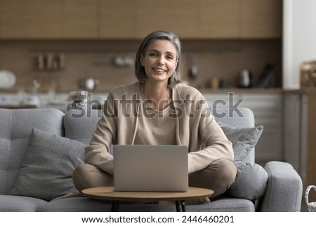 Carefree positive mature woman sitting on sofa in living room with laptop, looking at camera, posing, enjoy modern technology usage and on-line shopping, make order through electronic commerce website