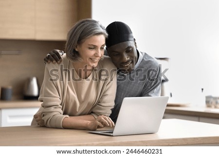 Loving carefree middle-aged spouses spend weekend time together at home, stand at kitchen table use laptop, look at screen, browse internet, read email, shopping on-line, discuss purchase, make order Royalty-Free Stock Photo #2446460231