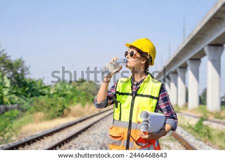 railway engineer or construction worker in hardhat and safety vest,holding paperwork is thirsty in the harsh sunlight,attractive caucasian female wears sunglasses drinking water while working outside
