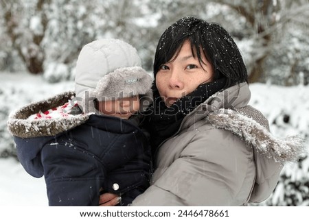 England, Europe - 12 18 2010 : Exterior photo view of a motehr and her new baby born kid boy child children in the snow snowy cold winter weather cold front in a park garden family portrait