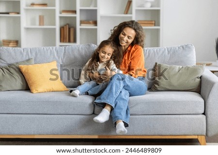 Smiling woman and little girl using cellphone, sitting on couch in living room, browsing internet, watching cartoon together, free space