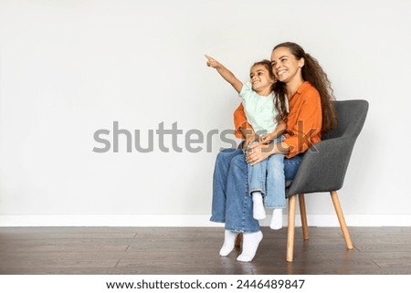 Girl pointing aside at free space while sitting on mother laps on chair, showing place for your advertisement over white wall, full length