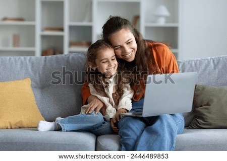 Happy caucasian mother and daughter using laptop and embracing, websurfing or watching cartoons online, sitting on sofa at home