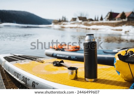 A thermos with a mug stands on a SUP against the background of a water landscape, a hot drink in cold weather, a tourist rafting on a SUP board. High quality photo