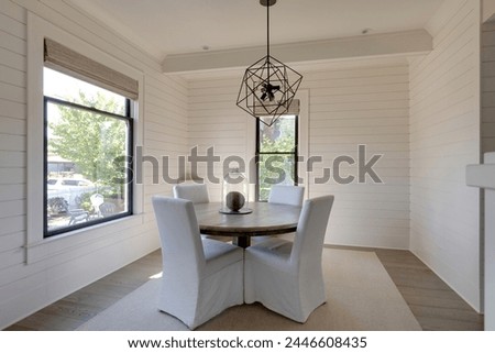 A cozy dining space with a stylish table setting, adorned by a chic decoration, beneath an elegant light fixture, framed by two bright windows. Pictured at a traditional cottage.