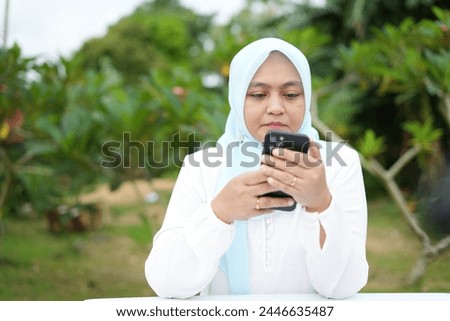 Asian woman in blue hijab is holding a cell phone with her facial expression while outdoors in the bright sun