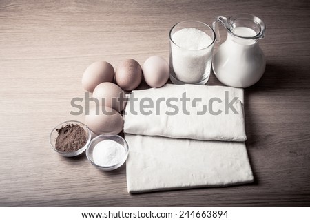 Milk, sugar, cocoa, puff dough, eggs and salt on a wooden board. Ingredients for pastry. Toned.