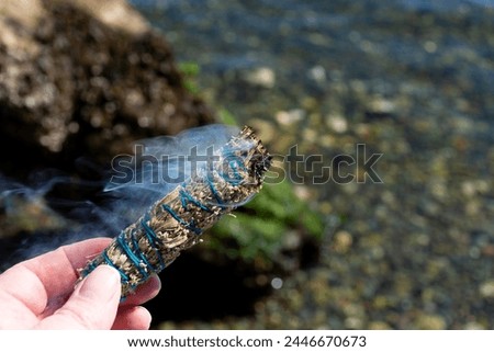 An image of a hand holding a burning white sage smudge stick in nature. 