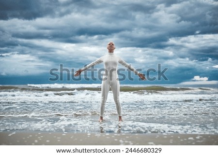 Happy hairless girl with alopecia in white futuristic suit stands with spread arms on beach bathed by ocean waves, performance of bald strong female artist symbolizing self acceptance