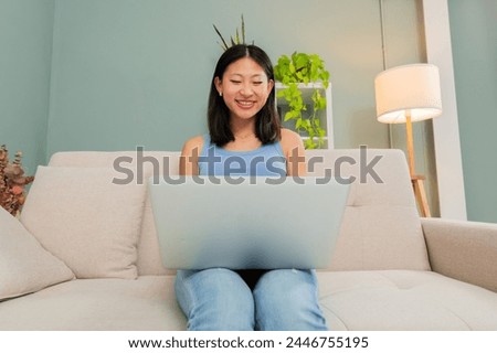 Joyful woman using the laptop sitting on the sofa and smiling minewhile shopping online. Font view of female sitting on the couch typing and surfing on social media with the computer in her apartment