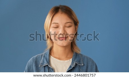 Close-up of young woman dressed in denim shirt stands with her eyes closed isolated on blue background in studio Royalty-Free Stock Photo #2446801757