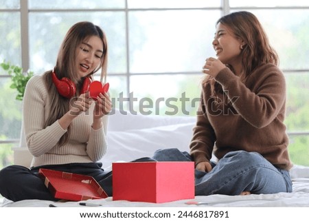 Asian female roommate sitting on bed, happy, surprised, receives a gift from friend, roommate reads card, opens gift, red headphones in apartment
