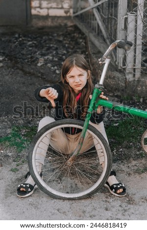 A little sad teenage girl, a dissatisfied upset child, showing a dislike, sits near an old bicycle with a broken, punctured wheel tire outdoors. Photography, portrait. Royalty-Free Stock Photo #2446818419