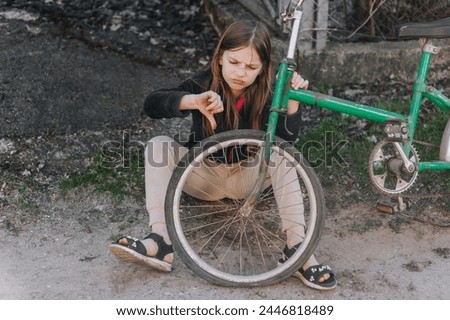 A small beautiful sad teenage girl, a dissatisfied upset child, showing a dislike, sits near an old bicycle with a broken, punctured wheel tire outdoors. Photography, portrait. Royalty-Free Stock Photo #2446818489