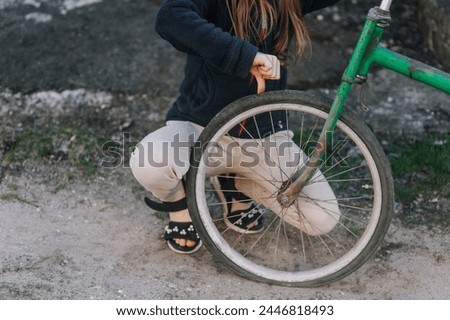 A teenage girl, a child, showing a dislike thumbs down sits near an old retro bicycle with a broken, punctured wheel tire outdoors. Close-up photography, portrait, lifestyle. Royalty-Free Stock Photo #2446818493