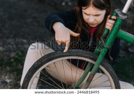 Little sad teenage girl, dissatisfied distressed child sitting near an old bicycle with a broken, punctured wheel tire outdoors. Photography, portrait. Royalty-Free Stock Photo #2446818495