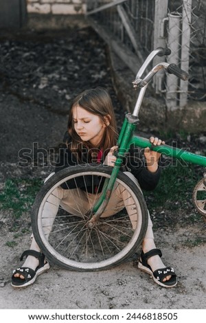 Little sad teenage girl, dissatisfied distressed child sitting near an old bicycle with a broken, punctured wheel tire outdoors. Photography, portrait. Royalty-Free Stock Photo #2446818505