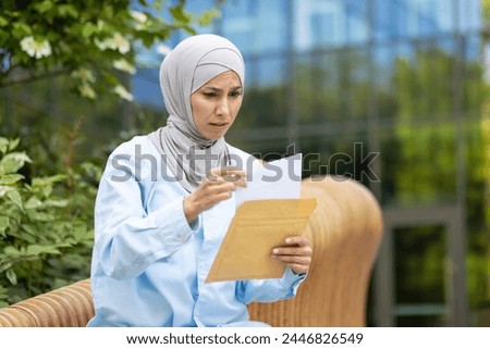 A worried Muslim woman wearing a hijab holding and reading a document or letter while sitting outside on a bench.