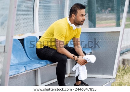 In the arbor of the city sports stadium, a bearded sportsman with headphones and a towel sits on a plastic chair, taking a well-deserved break on the outdoor athletic arena.  Royalty-Free Stock Photo #2446847835