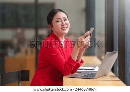 Asian businesswoman in a red jacket is sitting at a table with a laptop and a cell phone. She is smiling and she is enjoying her time