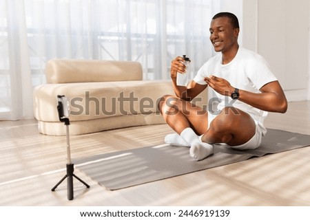 African American man smiles as he records a video on his phone mounted on a tripod, sitting on a yoga mat in a well-lit living room