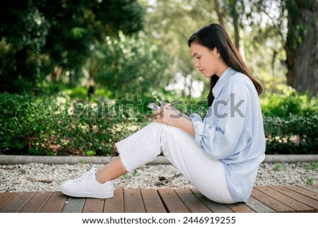 A young woman sits on the ground in a serene park setting, engaged with her smartphone, surrounded by lush greenery