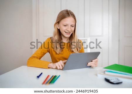 Cheerful teen girl engaged with a digital tablet at home, happy female teenager using modern gadget for study or leisure, sitting at desk indoors