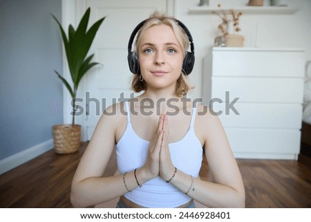 Image of young woman listens to yoga podcast for beginners, holds hands in namaste sign, wears headphones, meditates.