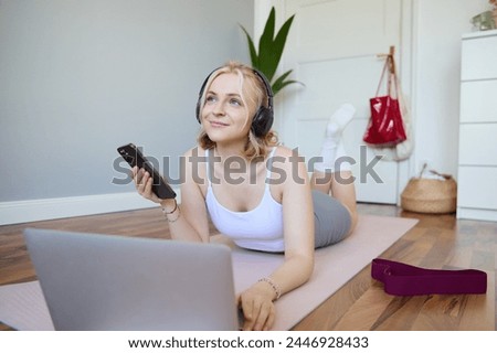 Portrait of happy, smiling sporty woman, doing workout at home, using laptop to watch workout videos in headphones, checking sports app on mobile phone.