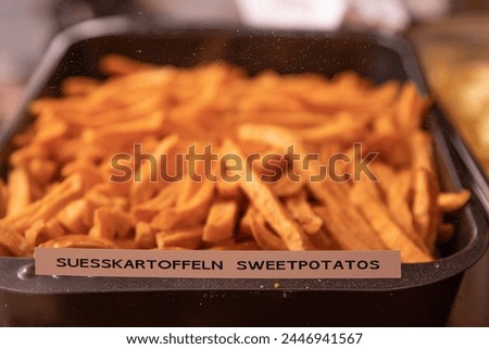 Yummy French fries in the metal tray with the tag Suesskartoffeln Sweetpotatos. Potato chips on the counter at the street market. Product Sign at market. Selective focus. Side view. Bokeh.