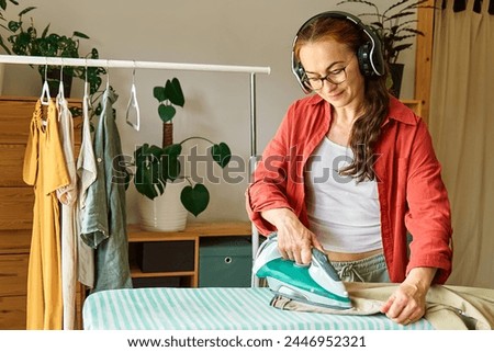 Happy middle aged woman in wireless headphones listening music, audiobook or podcast while ironing washed clothes on ironing board at home. Housewife daily routine. Housekeeping concept.