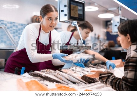 Female seller in apron standing near counter offering fresh fish hake to the buyer Royalty-Free Stock Photo #2446974153